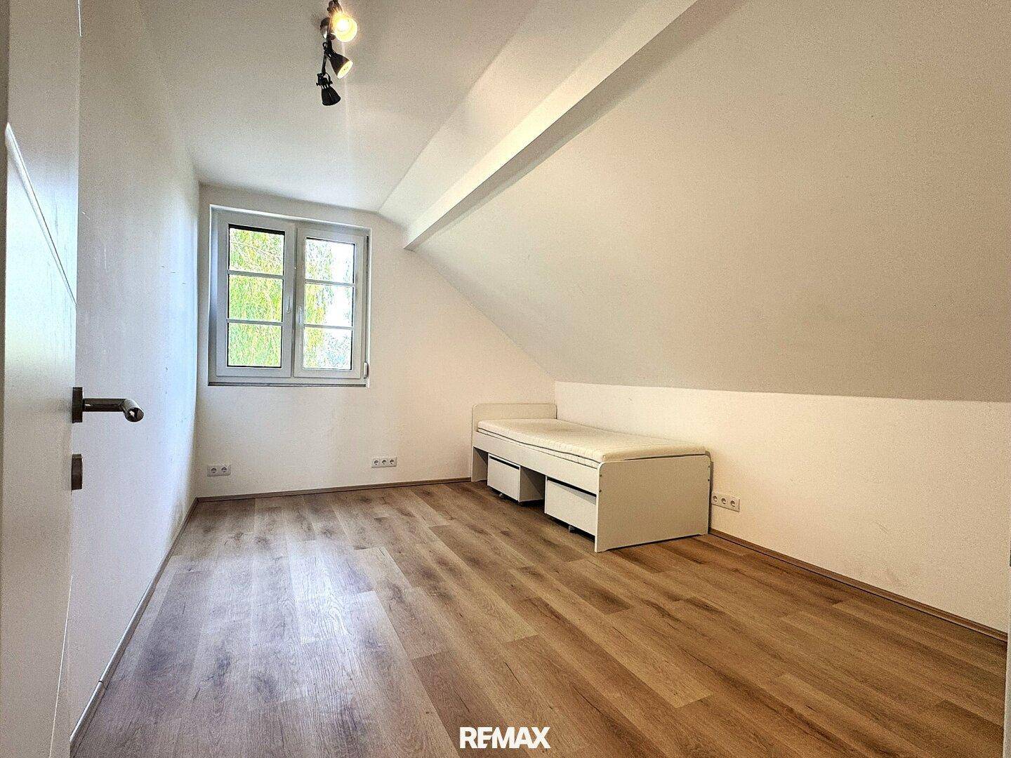 Helles Schlafzimmer im Dachgeschoss mit Laminatboden und einem Fenster mit Blick ins Grüne.