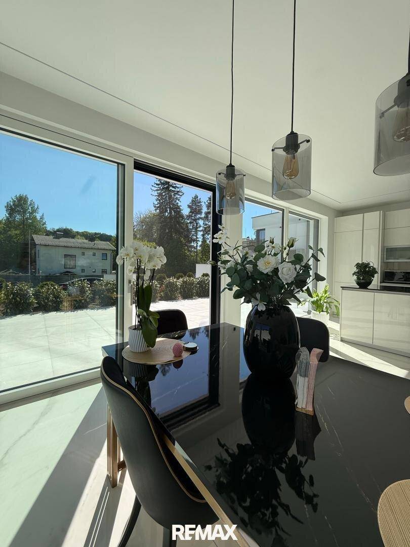 Essbereich mit Blick auf die moderne, weiße Küche und die umliegende Natur durch große Fenster.