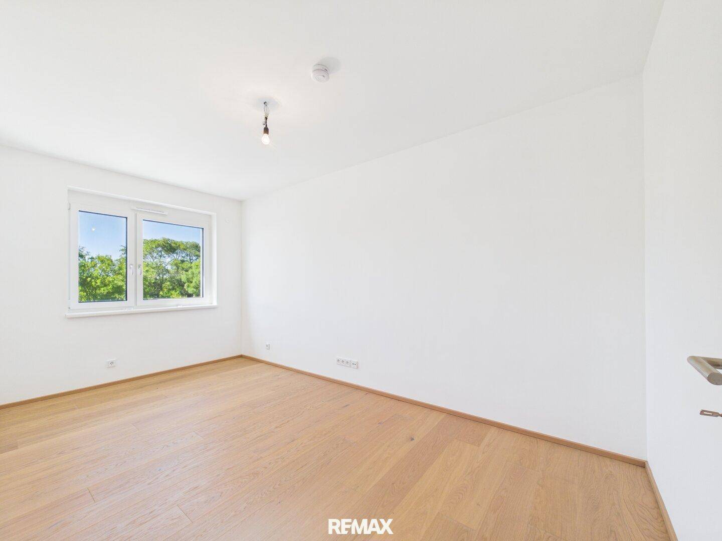 Geräumiges Schlafzimmer mit hellem Holzparkett und einem Fenster mit Blick ins Grüne.