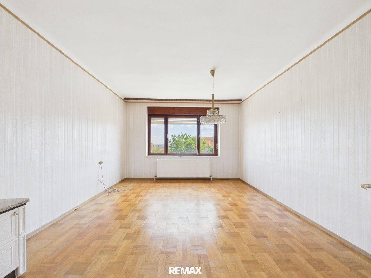 Geräumiges Wohnzimmer mit Parkettboden, großen Fenstern und Tapeten an den Wänden.