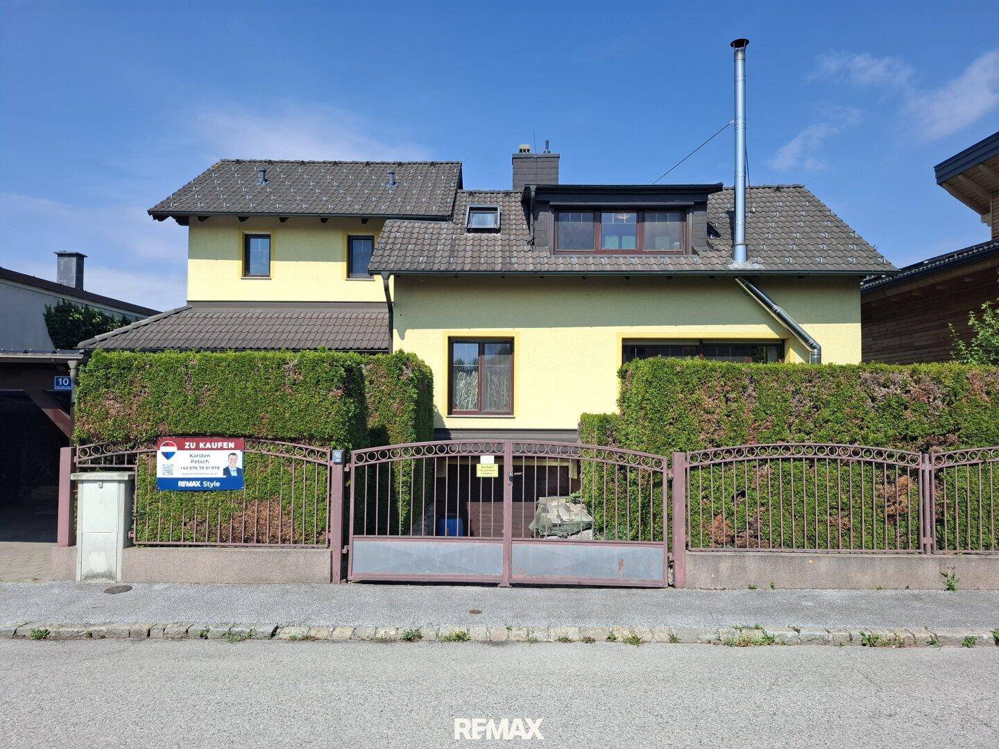 Vorderansicht des Hauses mit gelber Fassade, umzäuntem Vorgarten und Hecke.