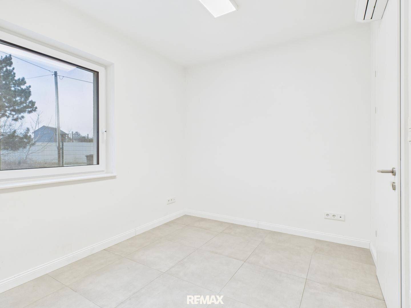 Leeres Zimmer mit hellen Fliesen, großem Fenster und Klimaanlage an der Wand.