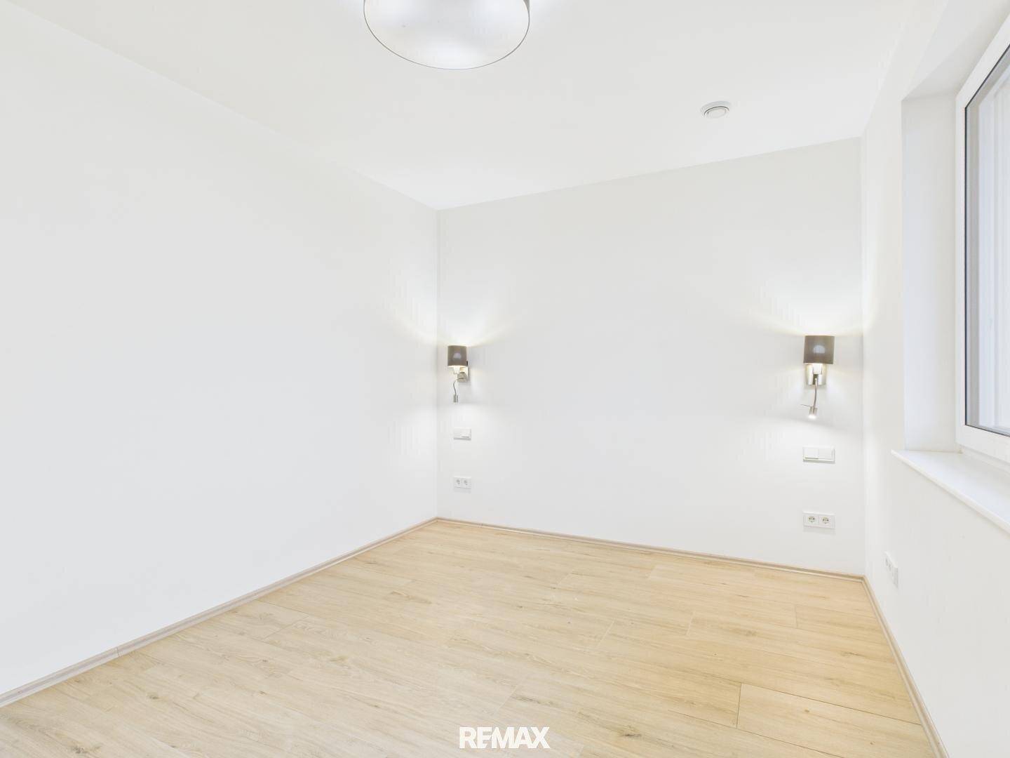 Leeres Zimmer mit hellem Laminatboden, weißen Wänden und zwei Wandleuchten.