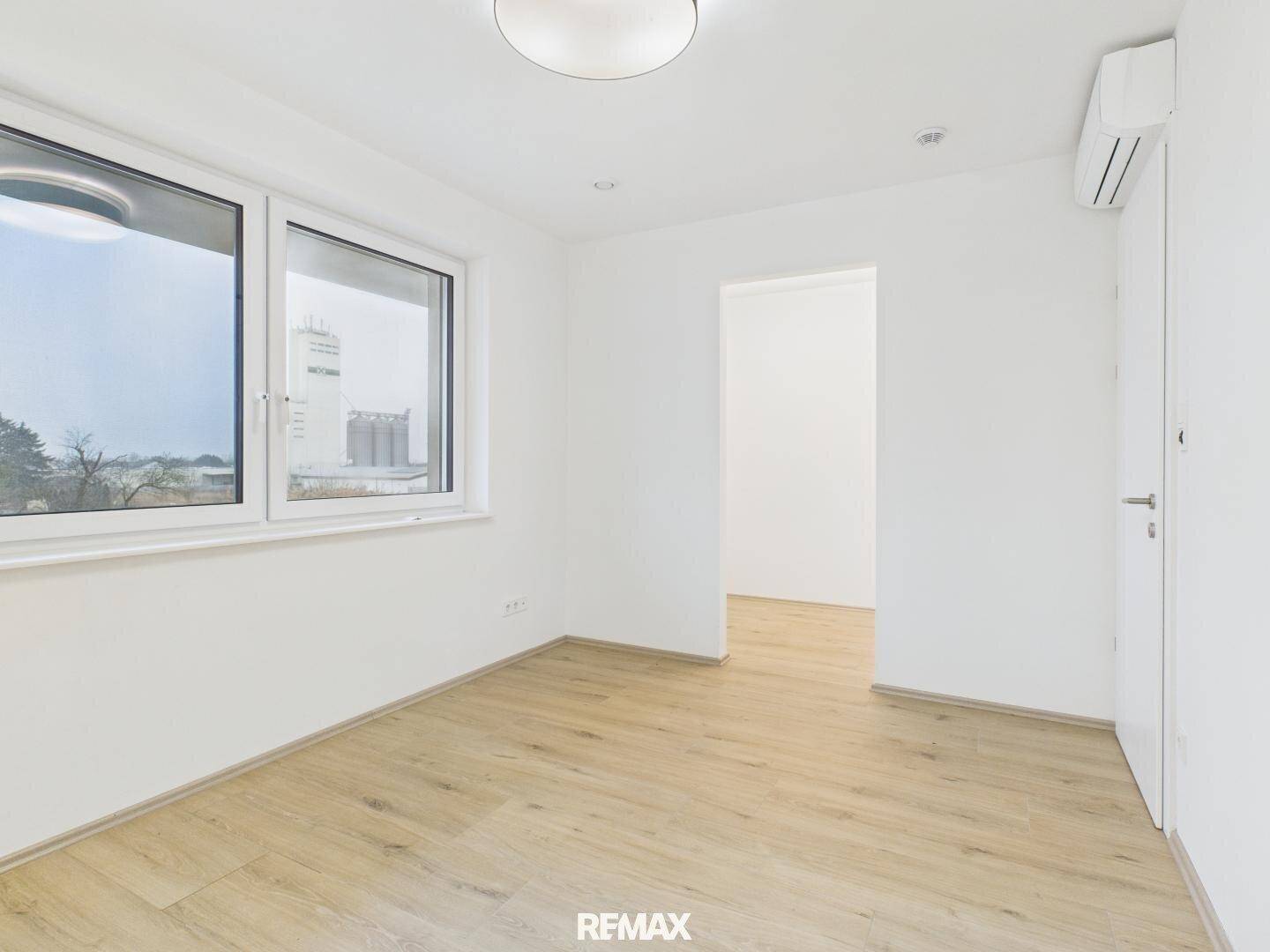 Helles Zimmer mit Laminatboden, großen Fenstern und Zugang zu einem weiteren Raum.