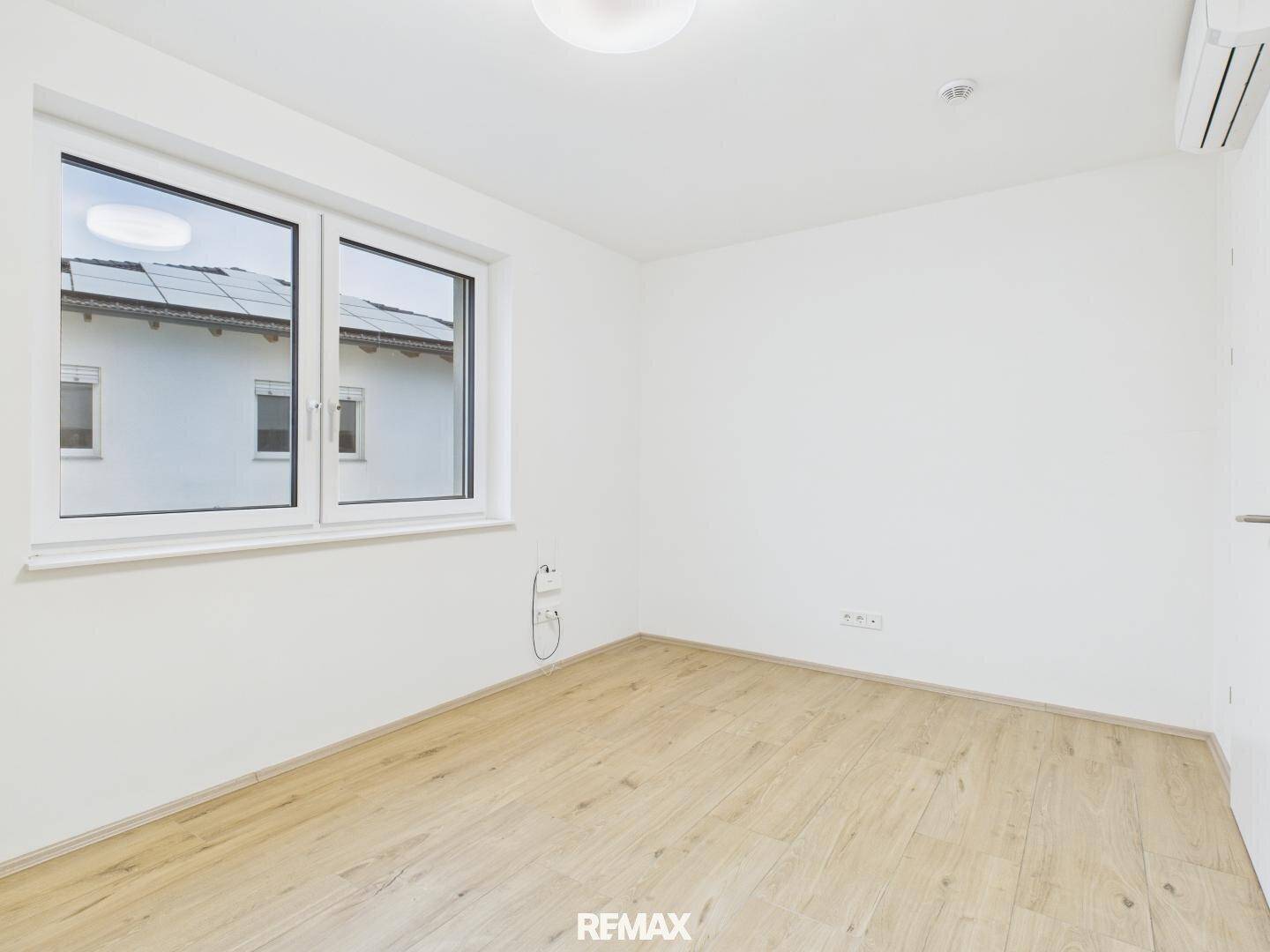 Leeres Zimmer mit Laminatboden, Fenster und Blick auf das Nachbargebäude mit Solaranlage.