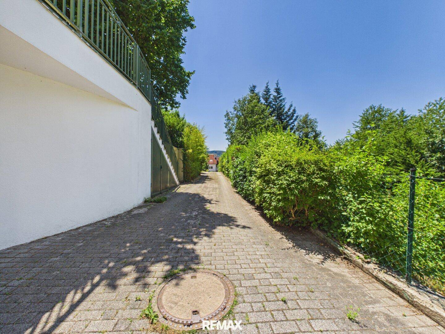 Gepflasterter Zufahrtsweg mit üppiger grüner Bepflanzung und Bäumen an einem sonnigen Tag.