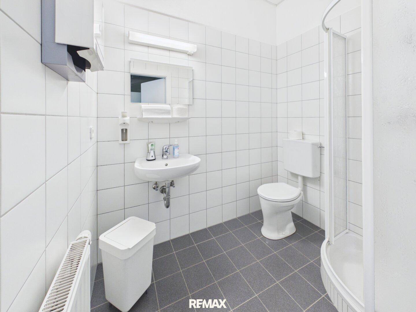 Funktionales Badezimmer mit Dusche, WC und Waschbecken, komplett gefliest in Weiß und Grau, sehr sauber.