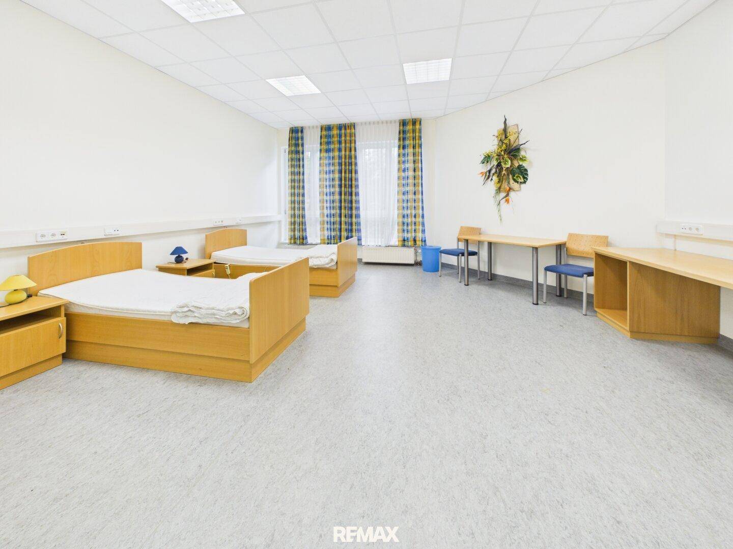 Helles Zimmer mit zwei Betten, Schreibtischen und bunten Vorhängen, geeignet als Patientenzimmer oder Mehrbettzimmer.