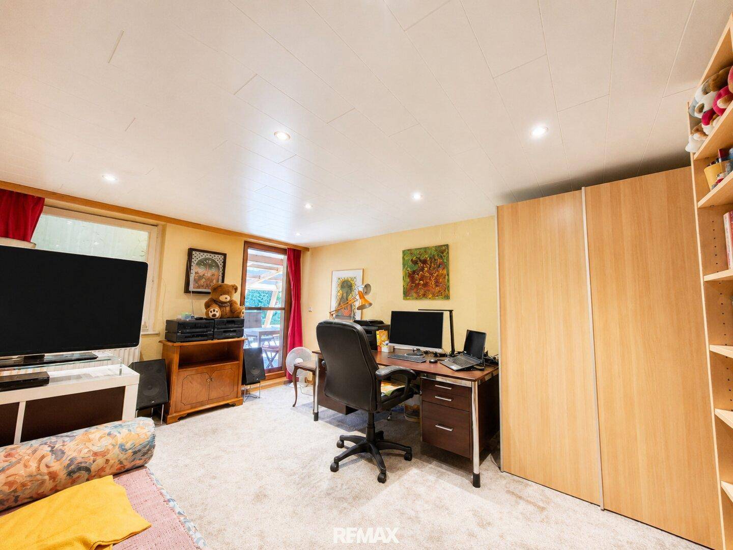 Geräumiges Arbeitszimmer mit Schreibtisch, Computer und großem Fernseher, ausgestattet mit Teppichboden.