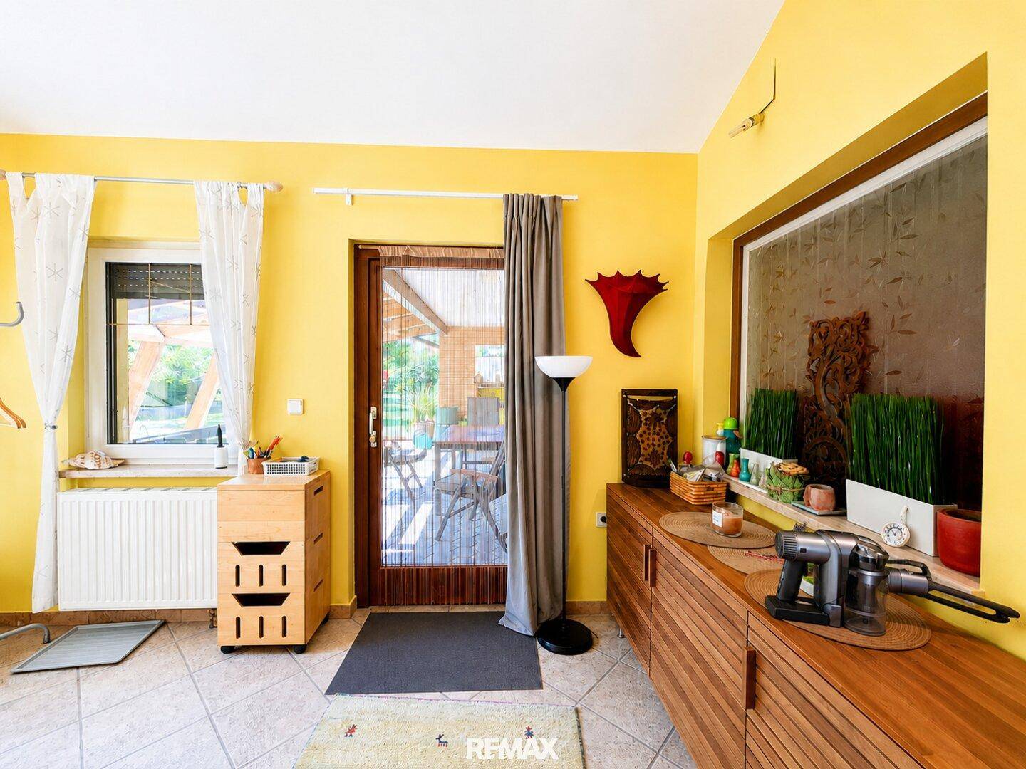Heller Eingangsbereich mit gelben Wänden, Fliesenboden und Zugang zu einer überdachten Terrasse.