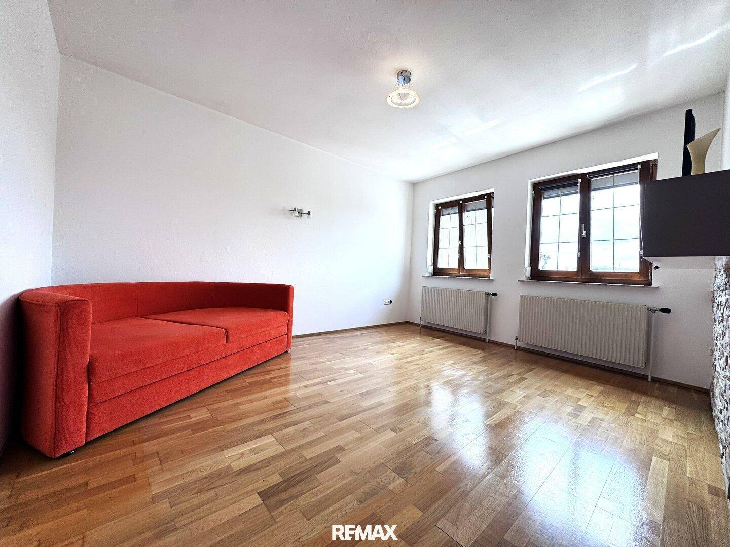 Gemütliches Zimmer mit Parkettboden, rotem Sofa und zwei Fenstern für Tageslicht.