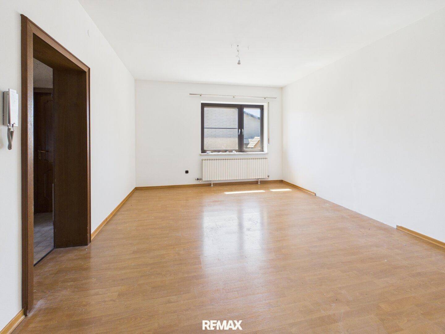 Helles, leeres Zimmer mit Holzboden und einem Fenster mit Blick ins Grüne.