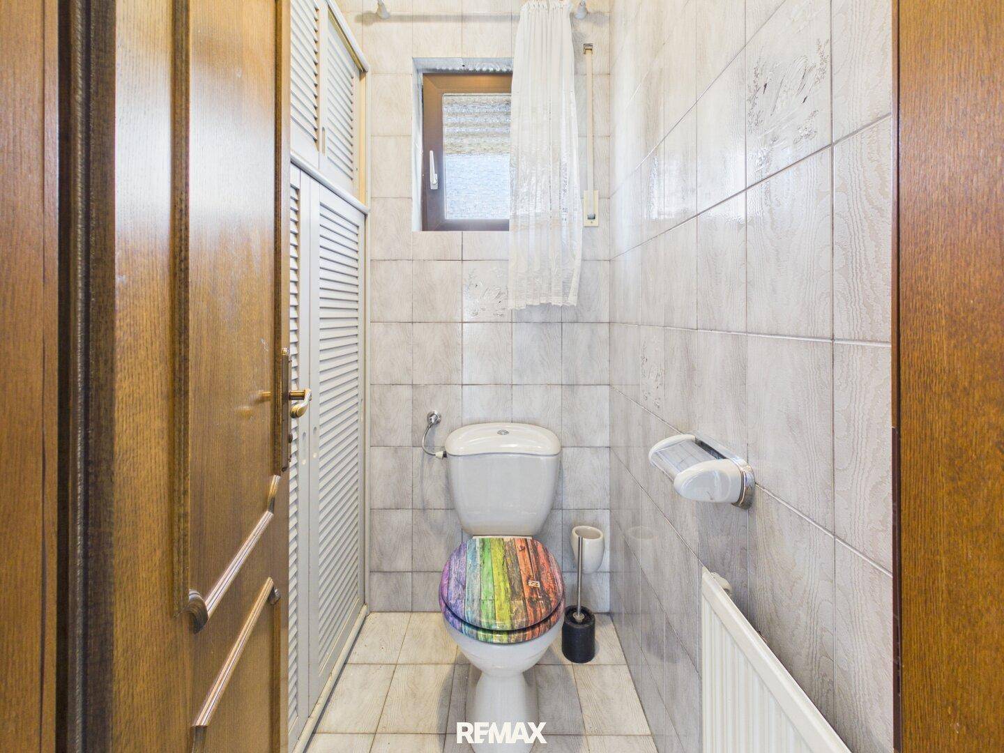 Kleines Gäste-WC mit Fliesen, Fenster und einer farbenfrohen Toilettenbrille.