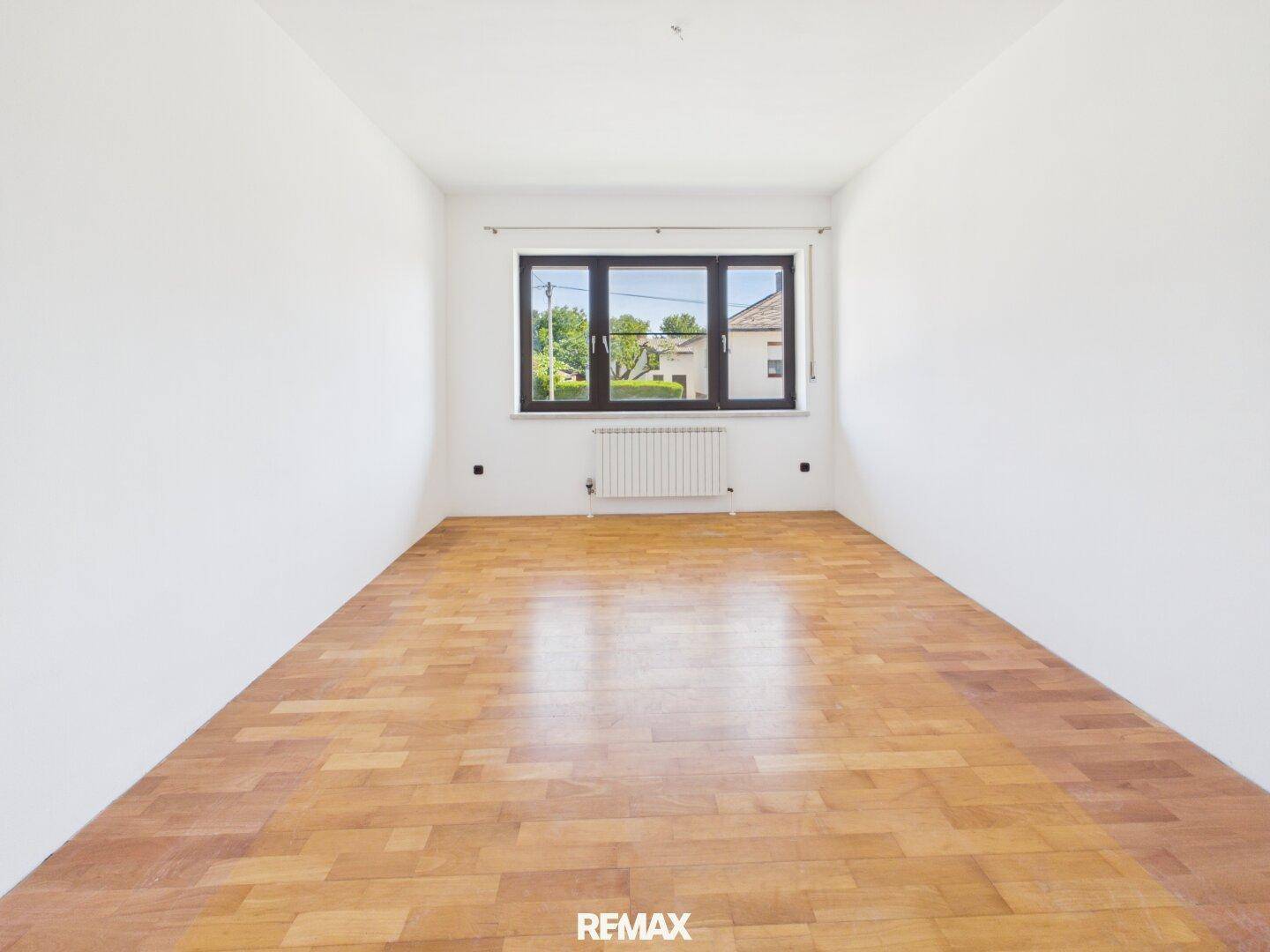 Freundliches, leeres Zimmer mit Parkettboden und großem Fenster für viel Tageslicht.