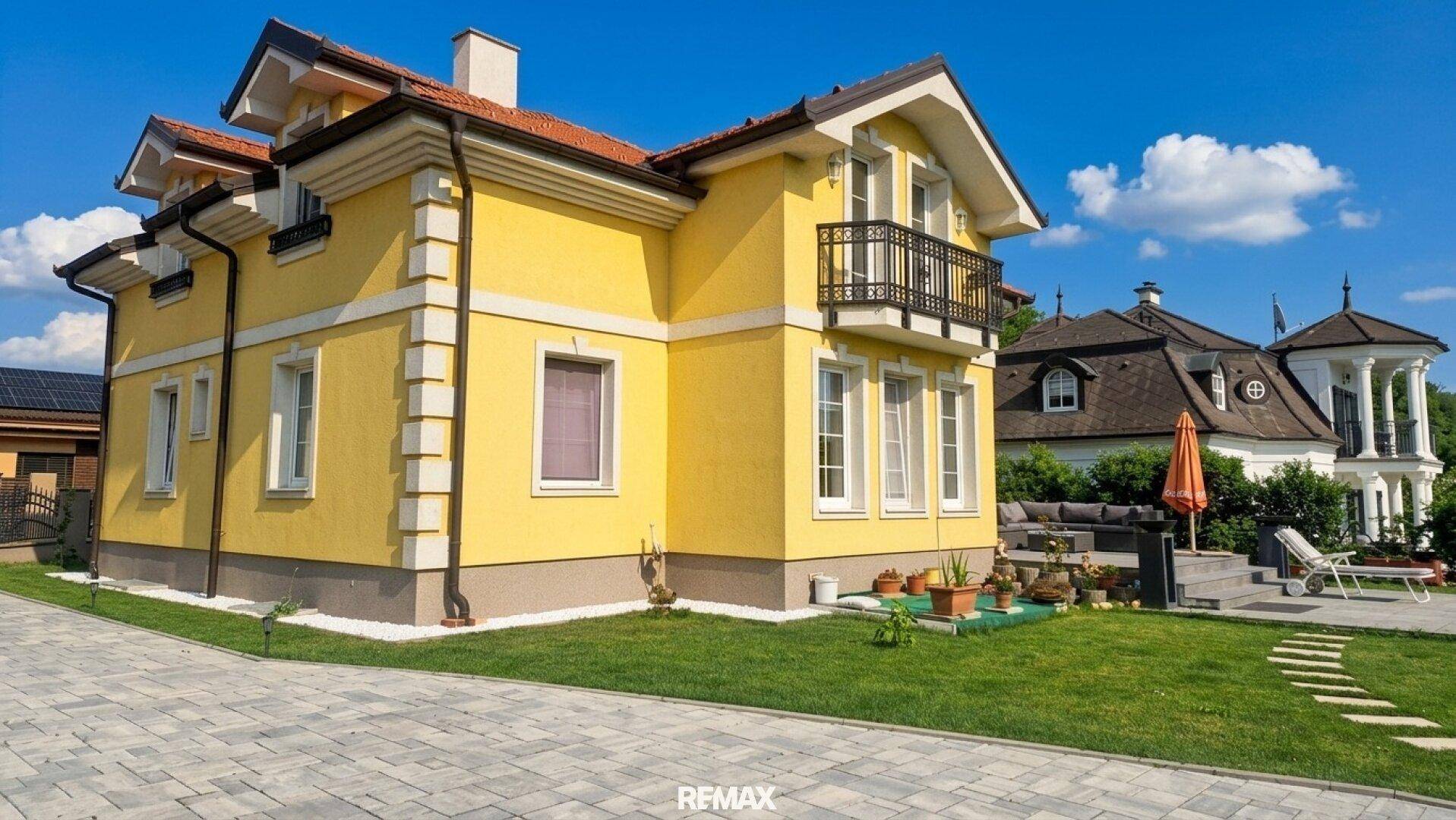 Seitenansicht der gelben Villa mit Balkon und gepflasterter Auffahrt.