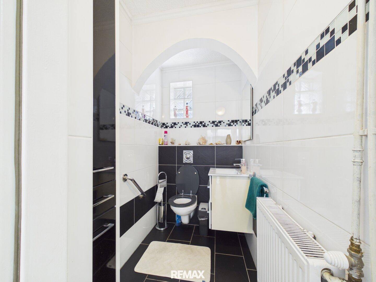 Kleines WC mit Toilette, Waschbecken und Haltegriffen, gefliest in Schwarz und Weiß.