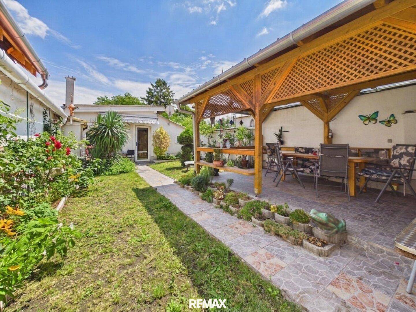 Gepflegter Garten mit Rasenfläche, Blumenbeeten und einer Holzpergola mit Sitzgelegenheiten.