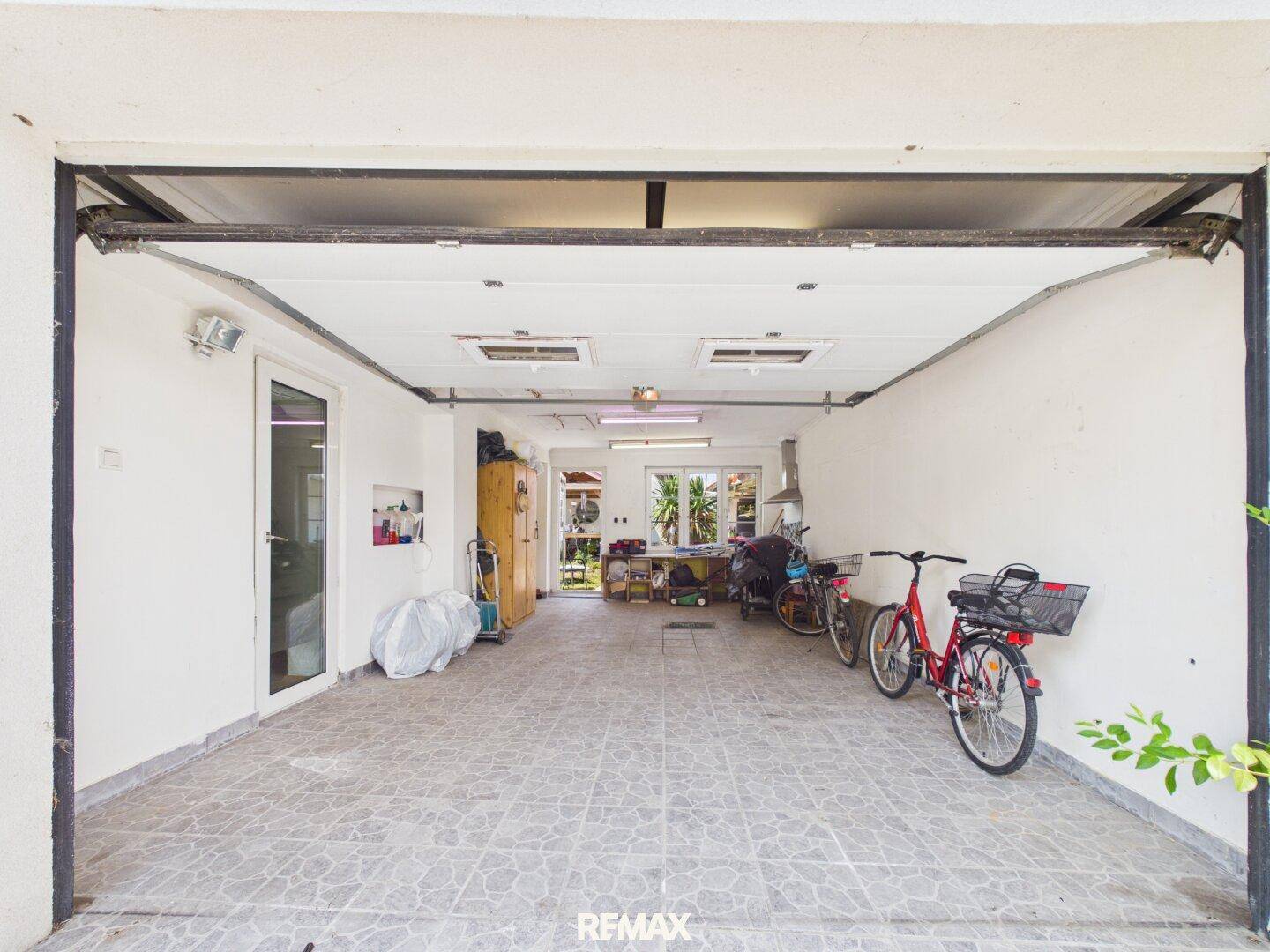 Geräumige Garage mit geöffnetem Tor, Fliesenboden und Fahrrädern, die im Inneren abgestellt sind.