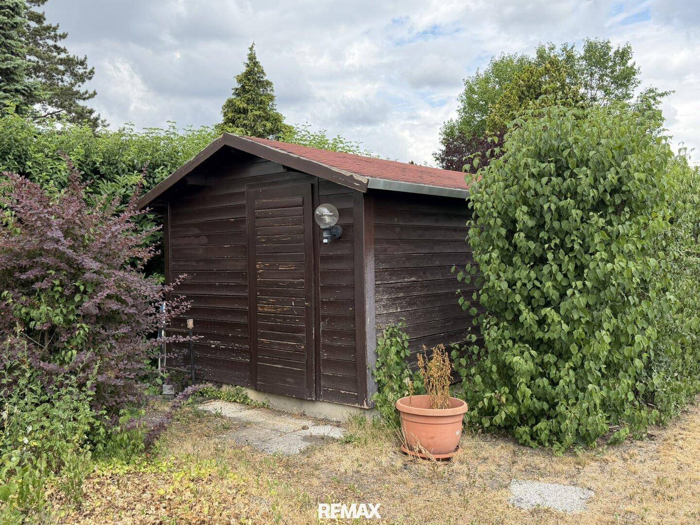 Kleiner Holzschuppen im Garten, umgeben von Sträuchern und Bäumen.