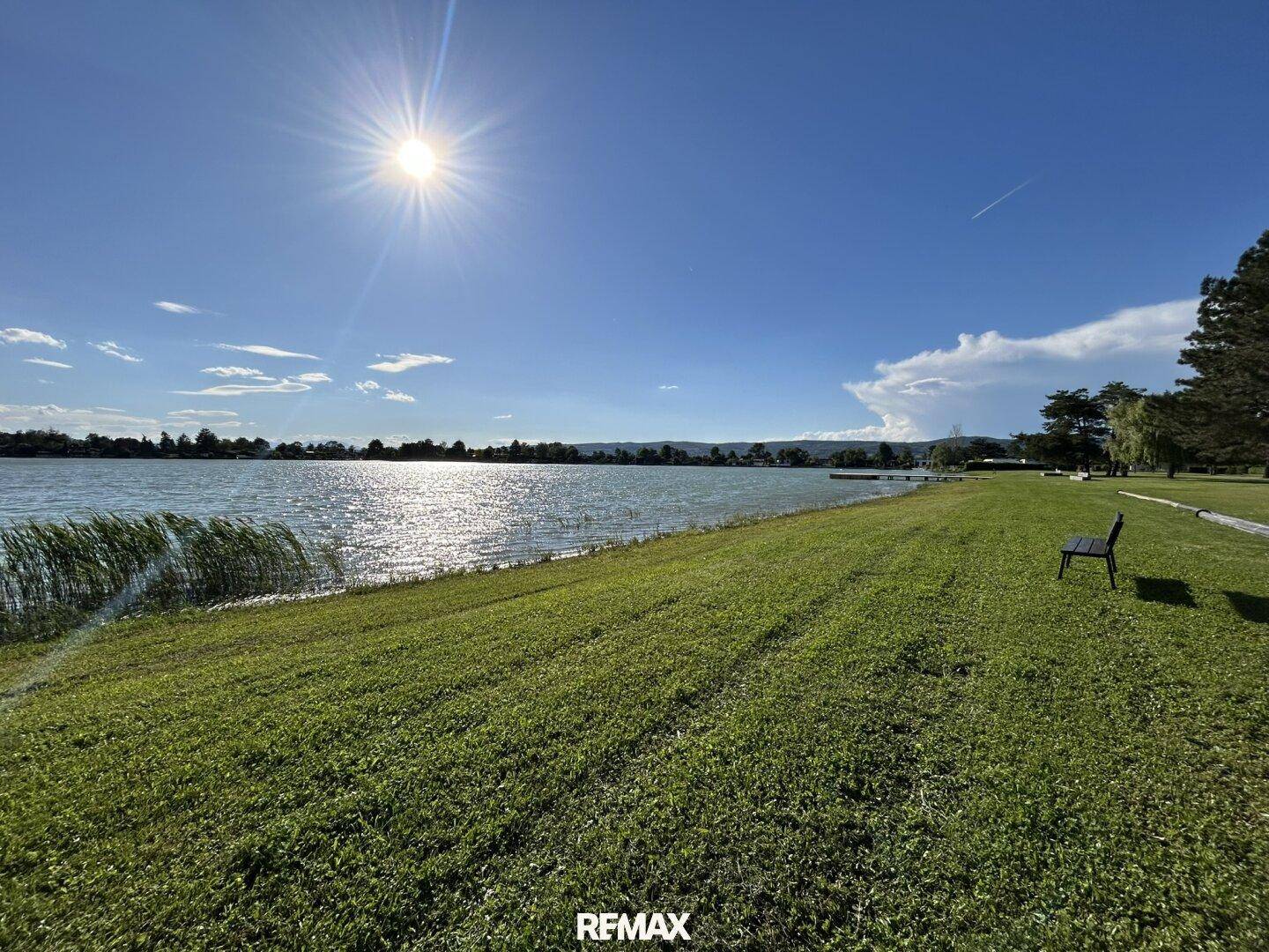Sonniger Blick auf einen ruhigen See mit grünen Ufern und blauem Himmel.