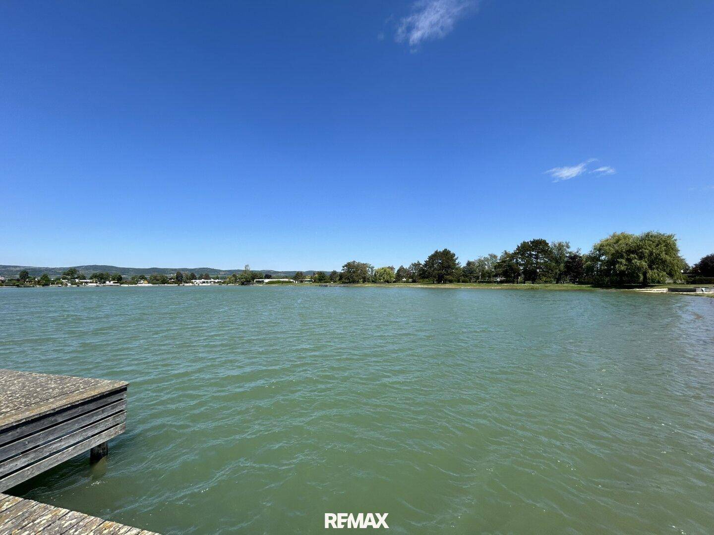 Weitläufiger Blick über den See mit einem Holzsteg im Vordergrund und bewaldeten Ufern.