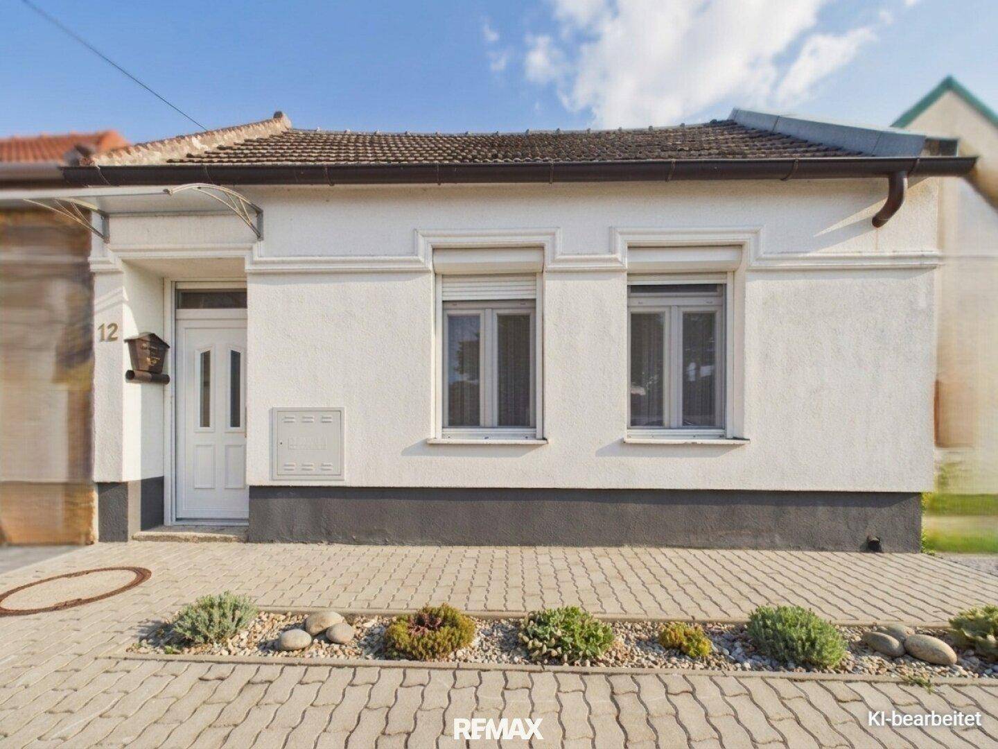 Weißes Einfamilienhaus mit dunklem Sockel und kleinen Fenstern, umgeben von einem gepflasterten Vorgarten.