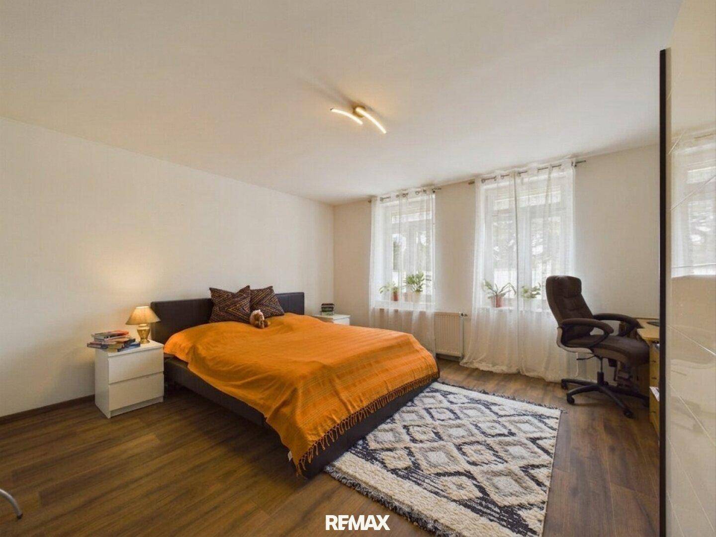 Helles Schlafzimmer mit Doppelbett, Schreibtisch und zwei Fenstern, ausgestattet mit Holzboden.