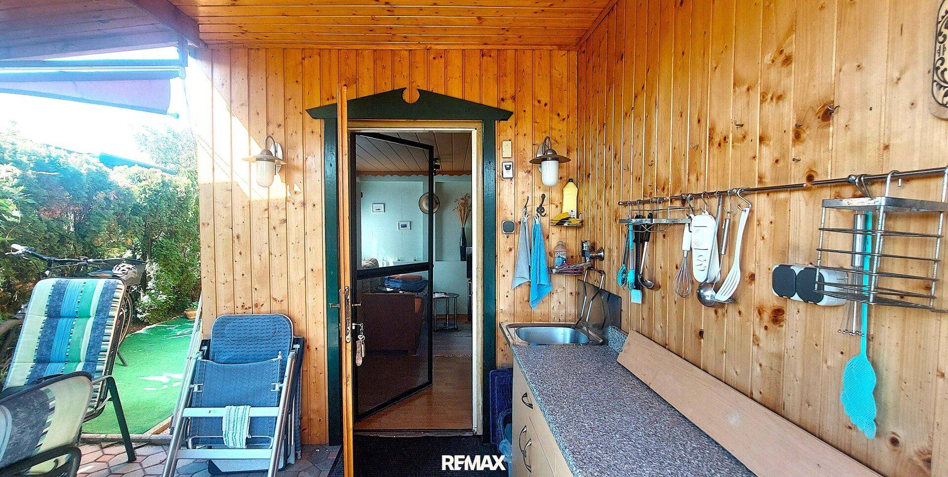 Überdachte Veranda mit Holzverkleidung, Außenspüle und Zugang zum Innenbereich, ideal für Mahlzeiten im Freien.