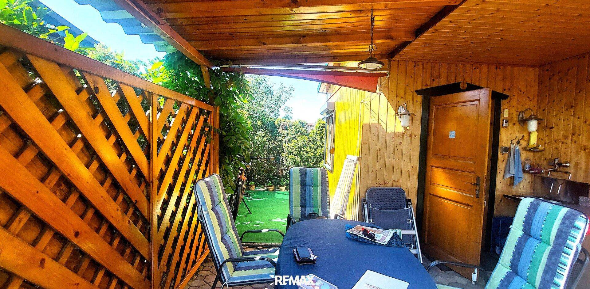 Überdachte Terrasse mit Holzzaun, Esstisch und Stühlen für entspannte Stunden im Freien, umgeben von Grün.