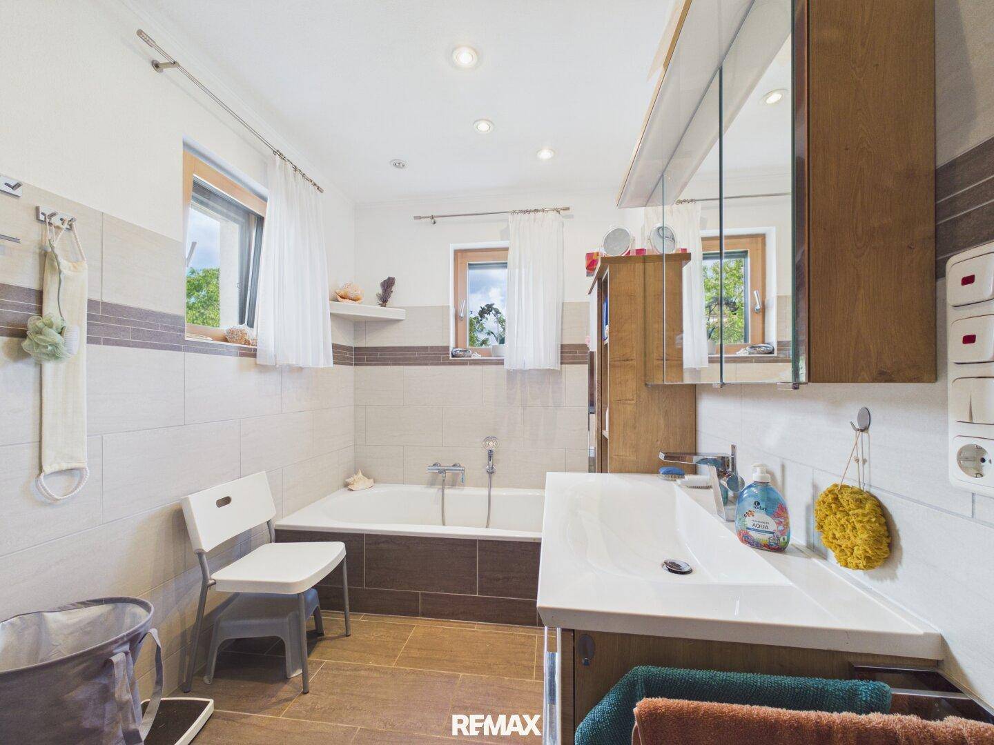 Modernes Badezimmer mit Badewanne, Einzelwaschbecken, Spiegelschrank und zwei Fenstern.