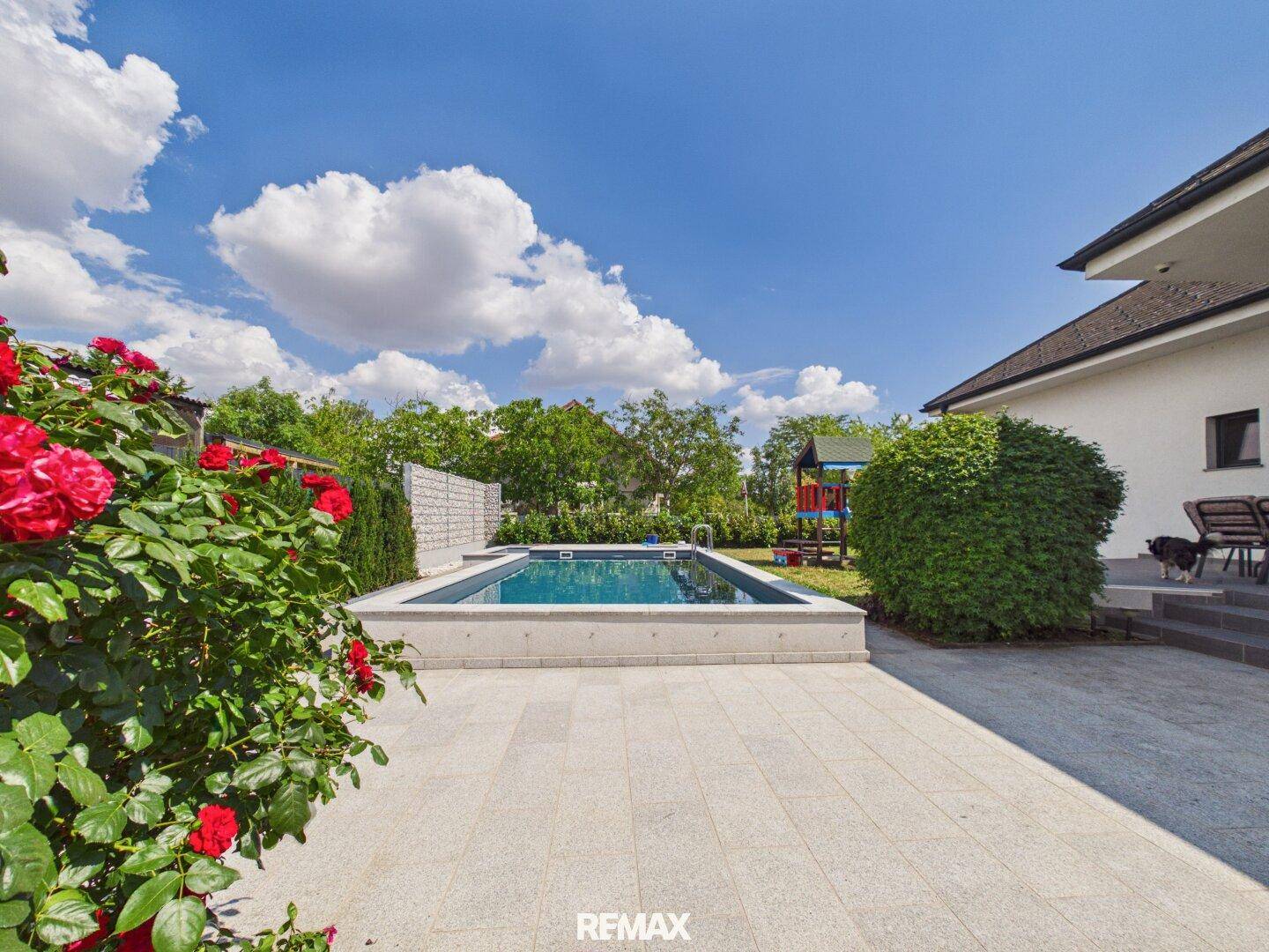Gepflegter Gartenbereich mit Pool, großer Terrasse und blühenden Rosenbüschen unter blauem Himmel.