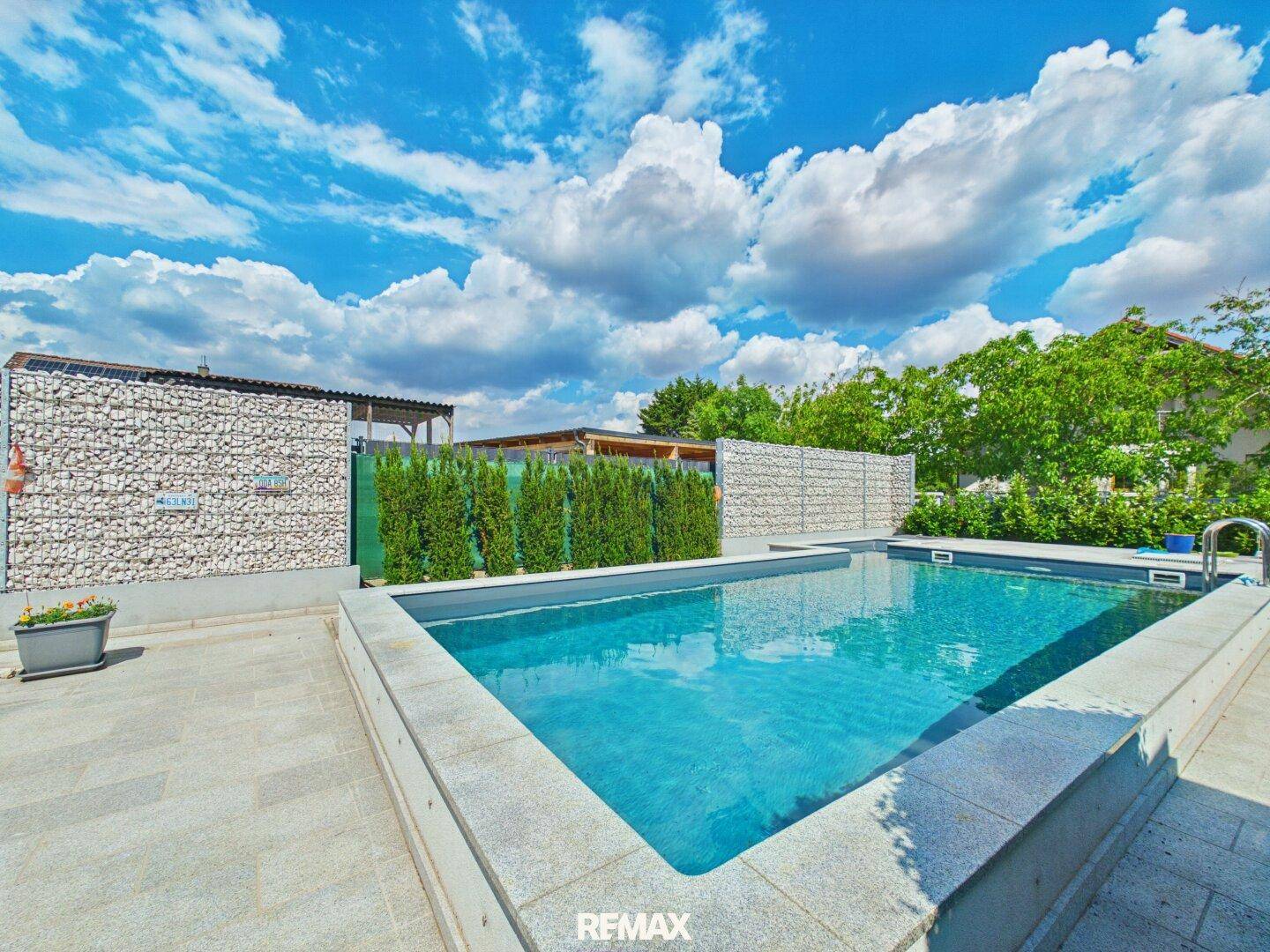 Außenpoolbereich mit gepflasterter Terrasse, Gabionenwänden und grünen Hecken.