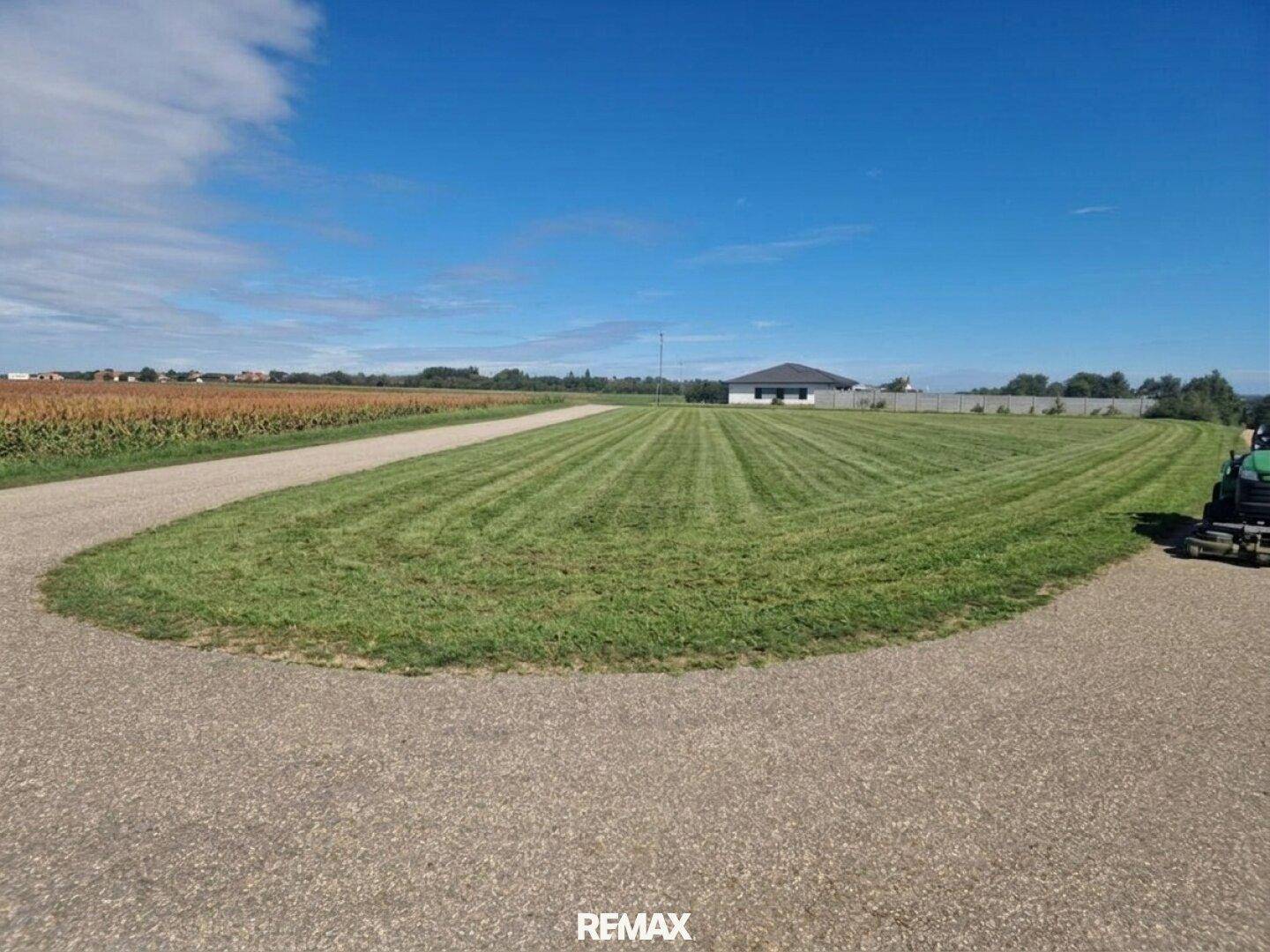 Gepflegte Rasenfläche mit geschwungenem Kiesweg und einem Neubau am Horizont unter blauem Himmel.