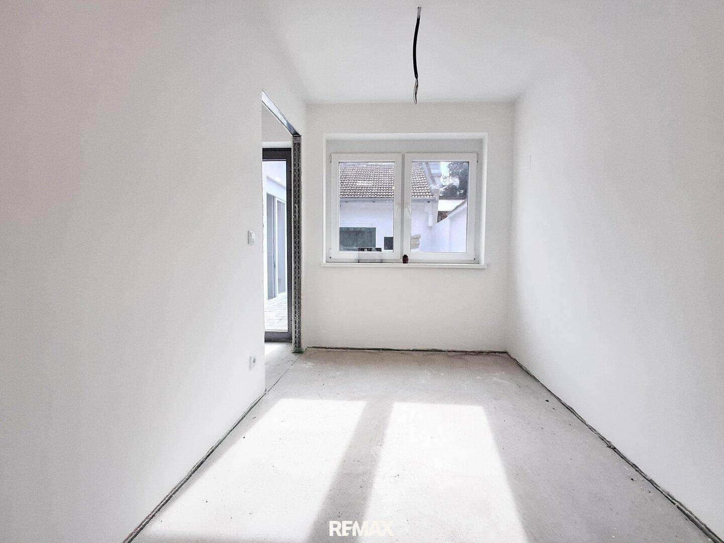 Leerer Raum mit weiß gestrichenen Wänden, Betonboden und einem Fenster, das Tageslicht hereinlässt.