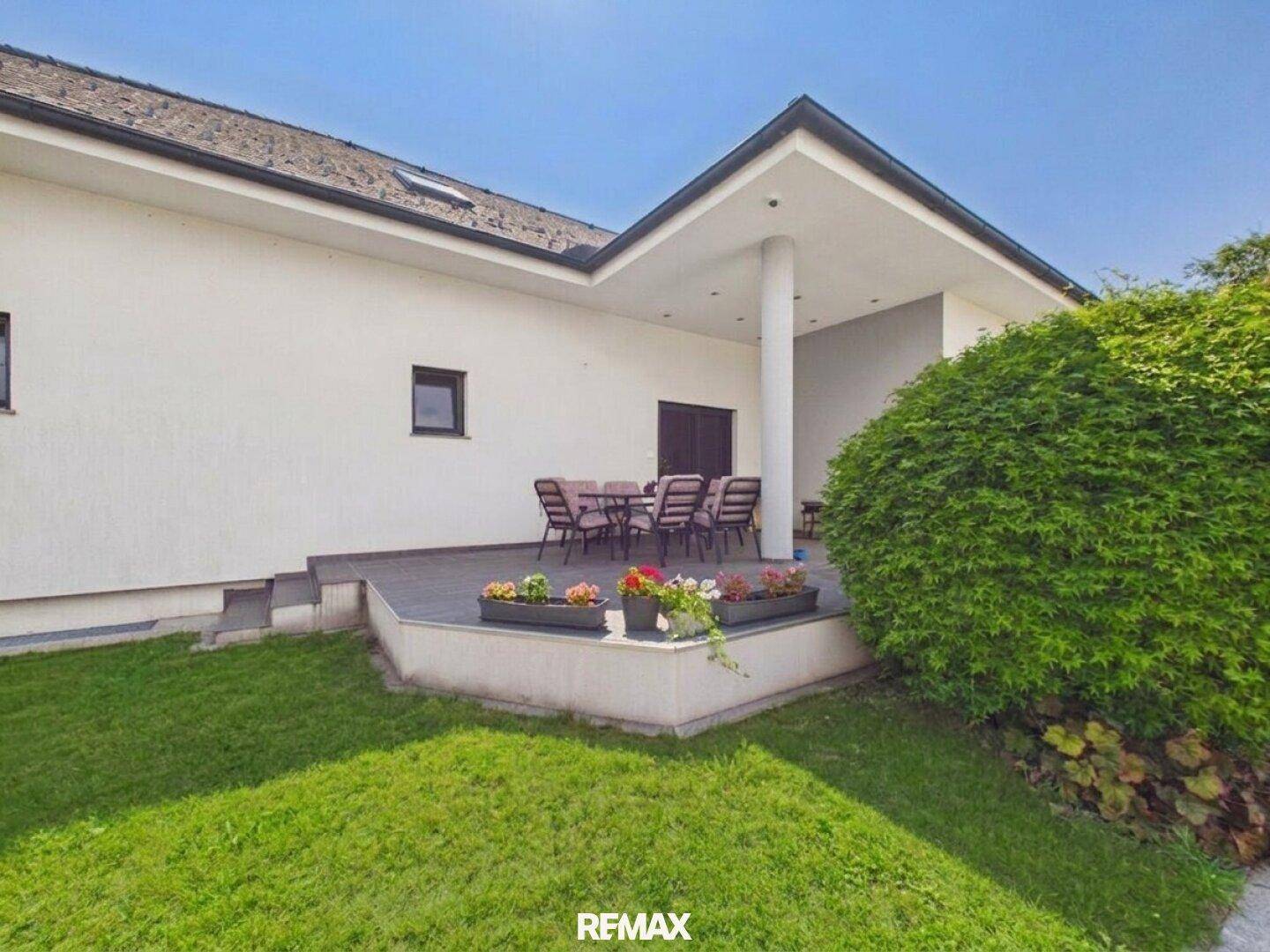 Überdachte Terrasse mit Gartenmöbeln, umgeben von Grünflächen und Sträuchern.
