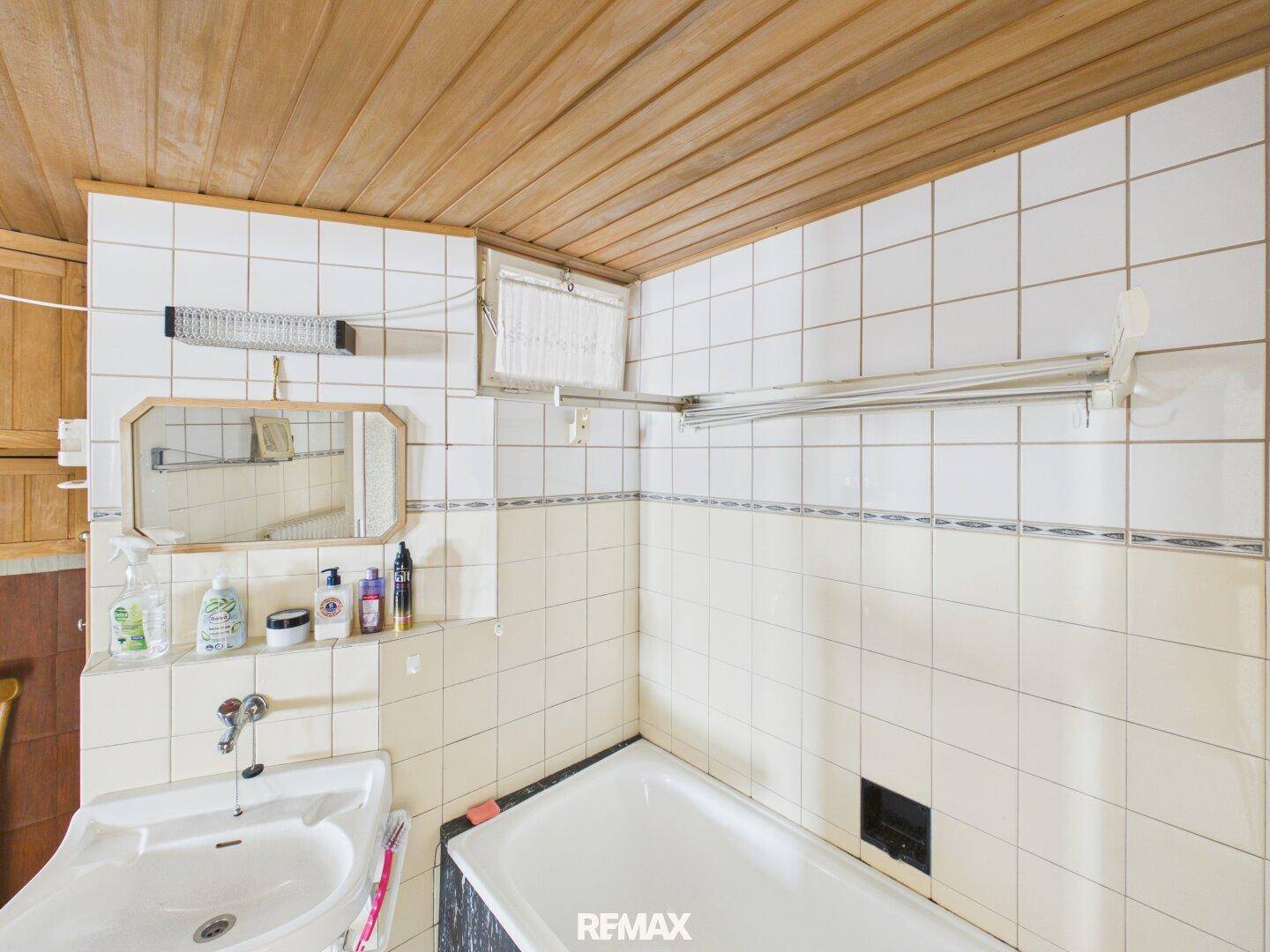Badezimmer mit Badewanne, Waschbecken und weißen Fliesen, mit Holzpaneeldecke.