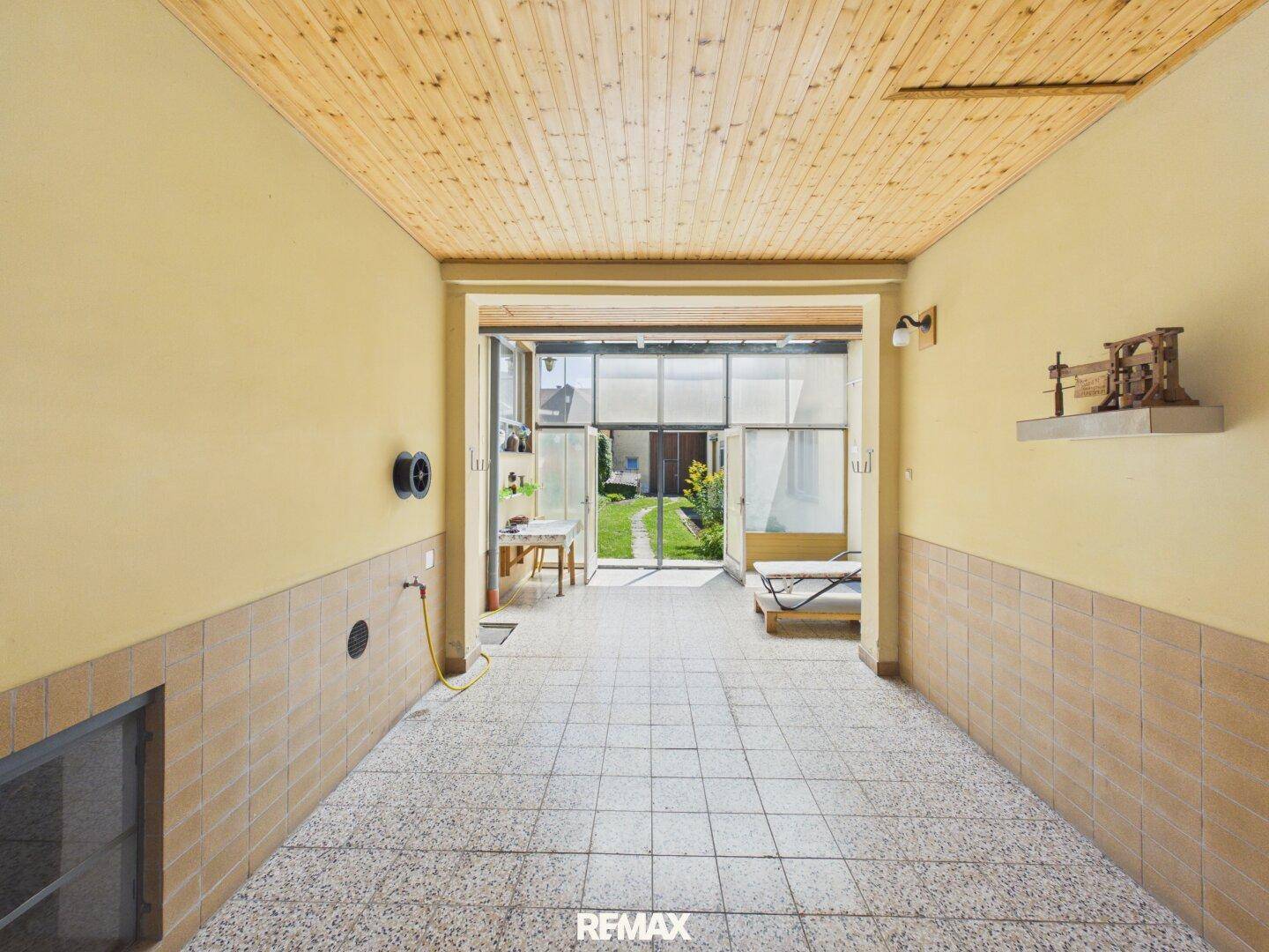 Durchgangsbereich mit Fliesenboden und Blick auf einen Garten, mit Holzpaneeldecke.