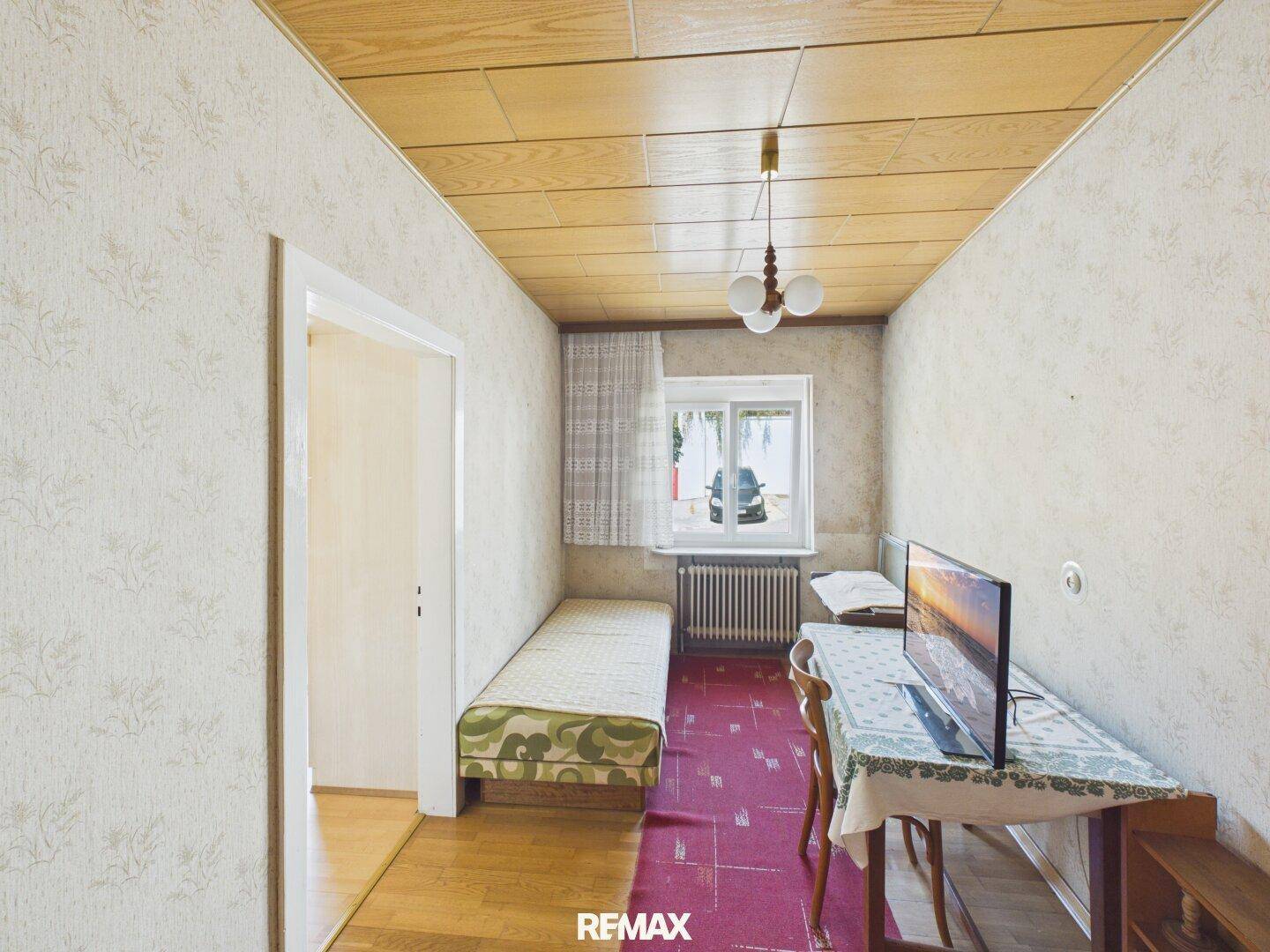 Kleines Zimmer mit Holzpaneeldecke, Einzelbett und einem Schreibtisch am Fenster.