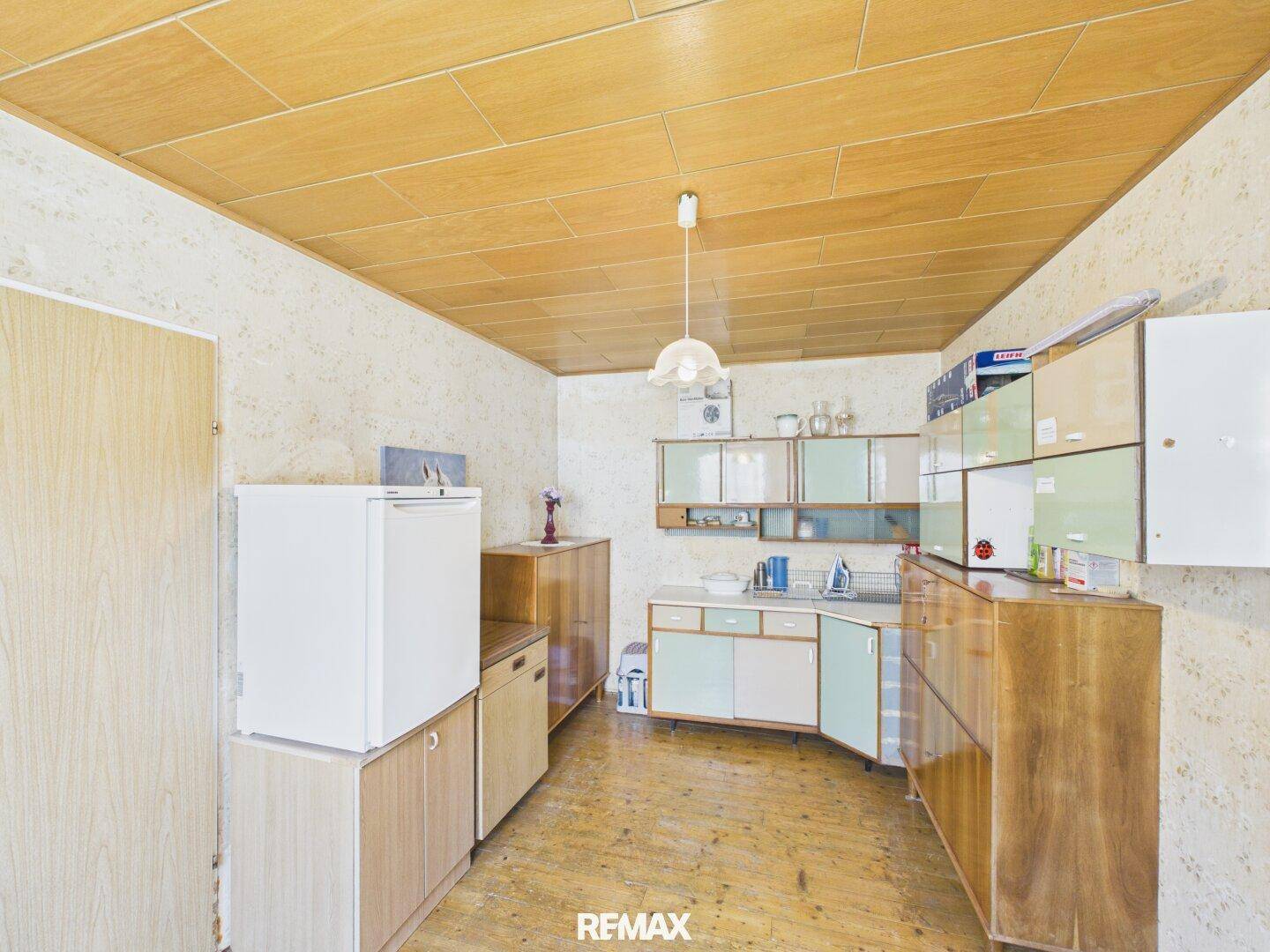 Einfache Küche mit älteren Schränken, Kühlschrank und Holzpaneeldecke.