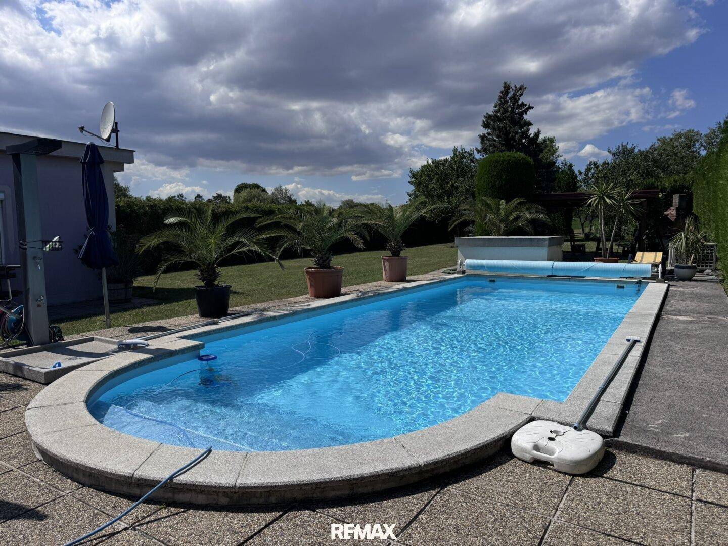 Ovaler Swimmingpool im Garten mit Steinumrandung und blauer Abdeckung unter bewölktem Himmel.