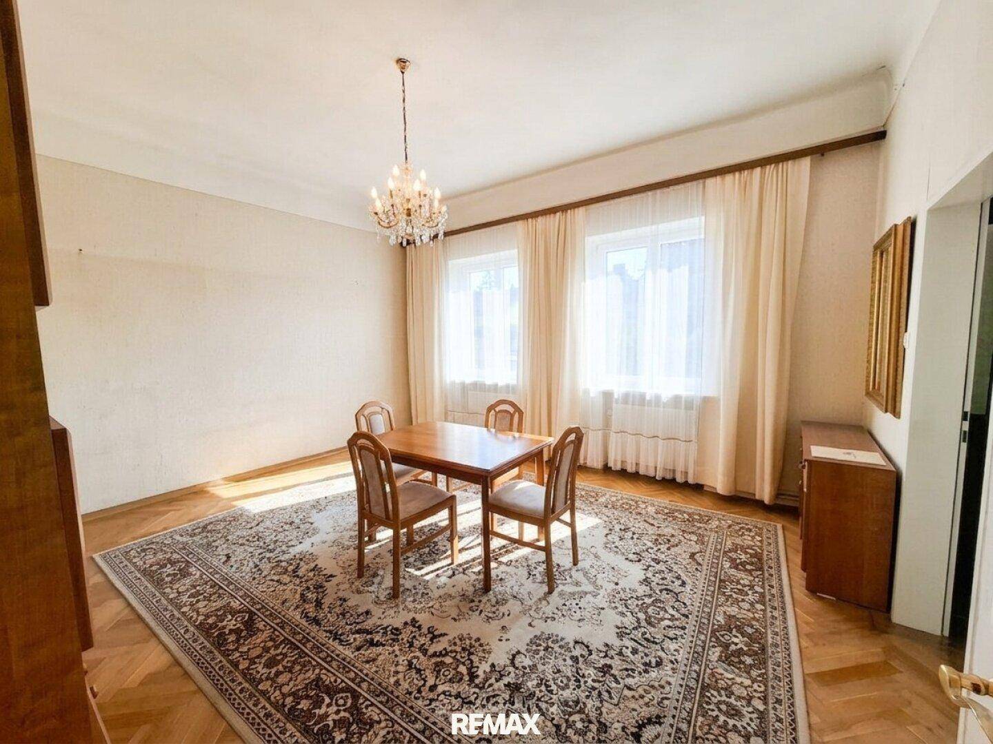 Helles Esszimmer mit Parkettboden, Kronleuchter und großem Fenster mit Vorhängen.