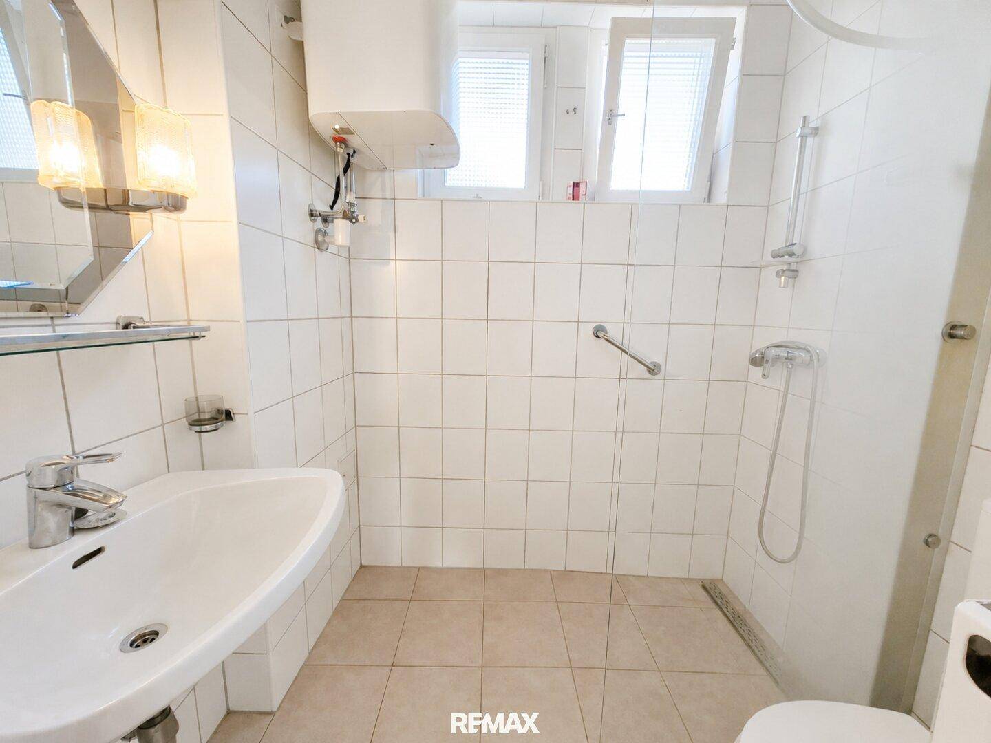 Funktionales Badezimmer mit Dusche, Waschbecken, weißen Fliesen und Haltegriffen.