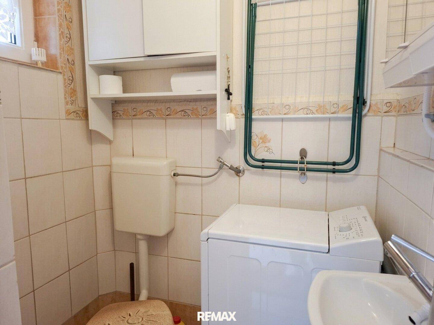 Kleiner Raum mit Toilette, Waschmaschine und Waschbecken, gefliest in hellen Tönen.