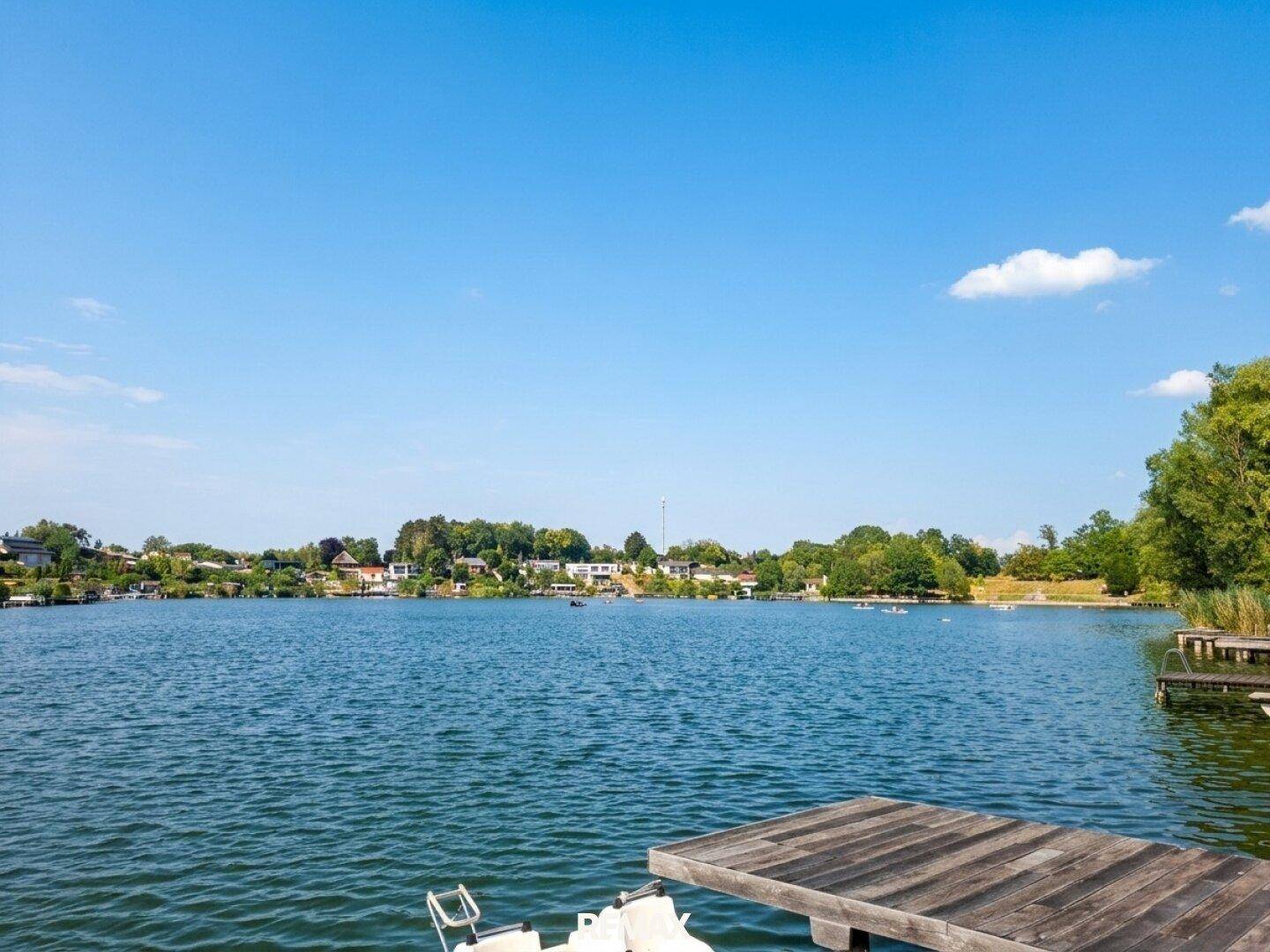 Weitläufiger Blick über den ruhigen See mit bewaldeten Ufern und blauem Himmel.