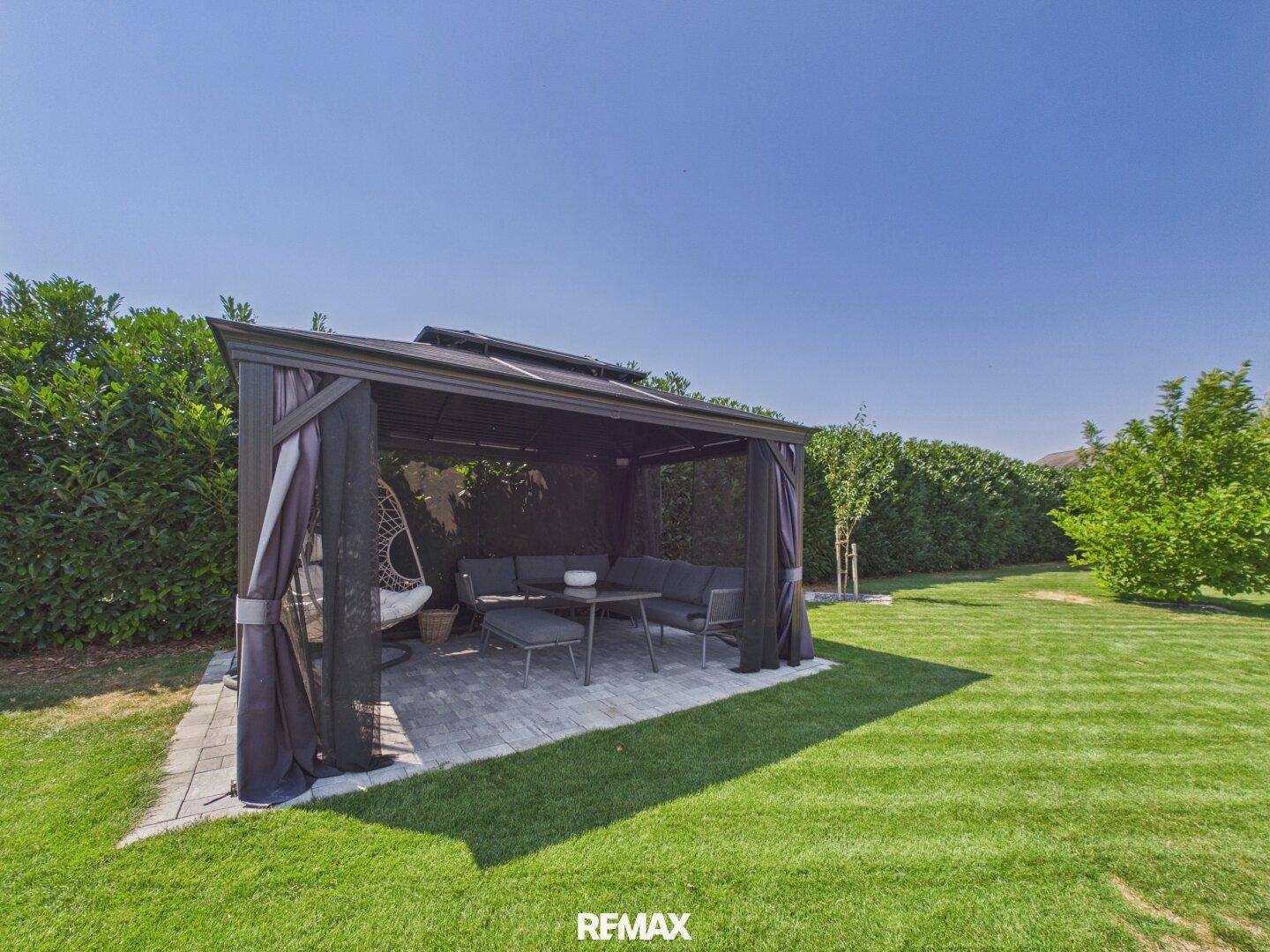 Gemütlicher Pavillon im Garten mit Lounge-Möbeln und umgeben von hohen Hecken.