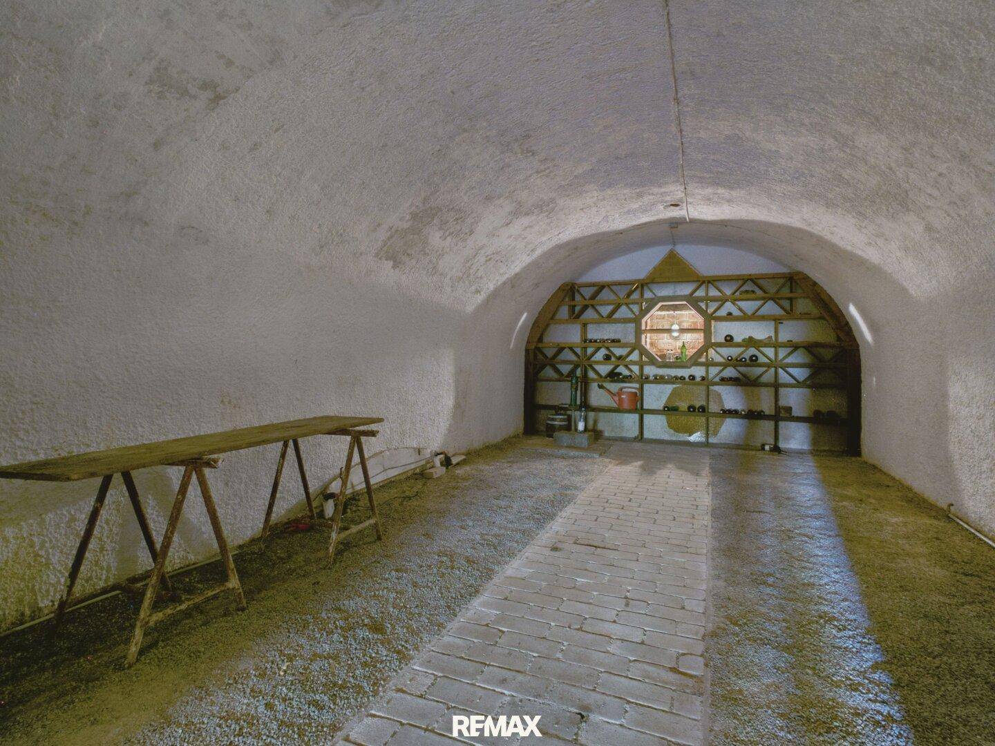 Langer Gewölbekeller mit Ziegelweg und Regalen zur Weinlagerung am Ende des Raumes.
