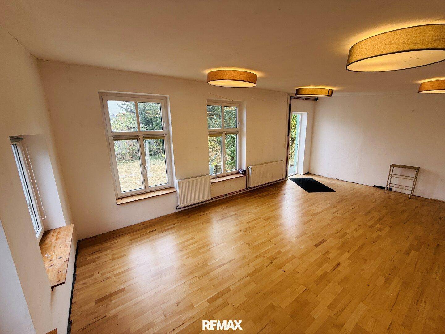 Großer, leerer Raum mit Holzfußboden, mehreren Fenstern und modernen Deckenleuchten.