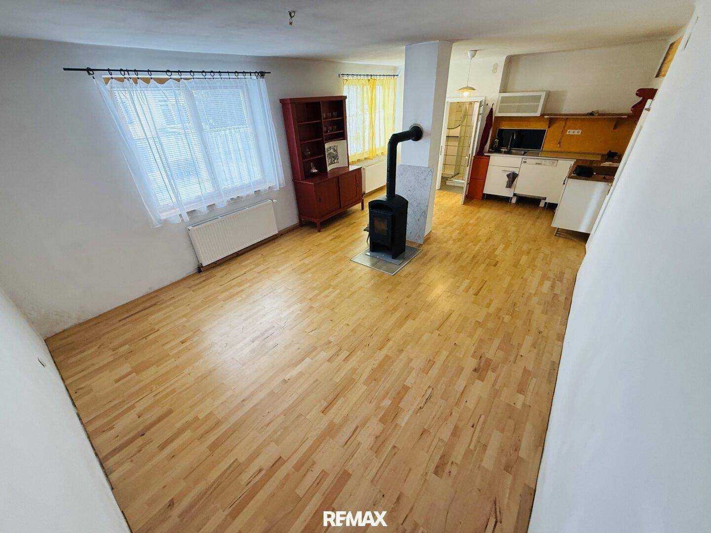 Wohnbereich mit Holzfußboden, einem Holzofen, Fenster und einem angrenzenden Küchenbereich.