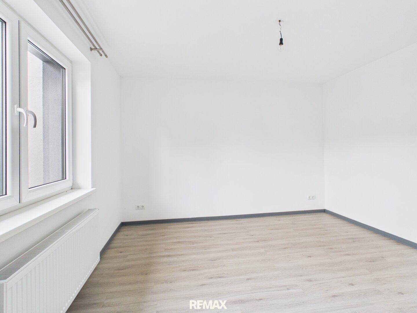 Leerer, heller Raum mit Laminatboden und einem großen Fenster, bereit zur Gestaltung.