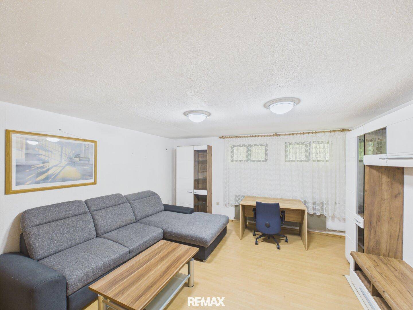 Helles Wohnzimmer mit grauem Ecksofa, Schreibtisch und Laminatboden.